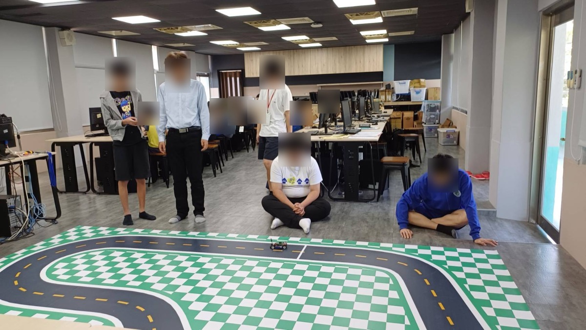 教室裡的實跑：學生和老師圍著地上的 TopRacer 賽道，車正在跑。臉部已做模糊處理。